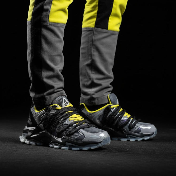 Gray and black sneakers with yellow accents worn by a person on a black background